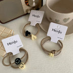 Three sets of beige hair ties with charms (smiley faces, hearts, flowers) are arranged on a table with a radio and cup.
