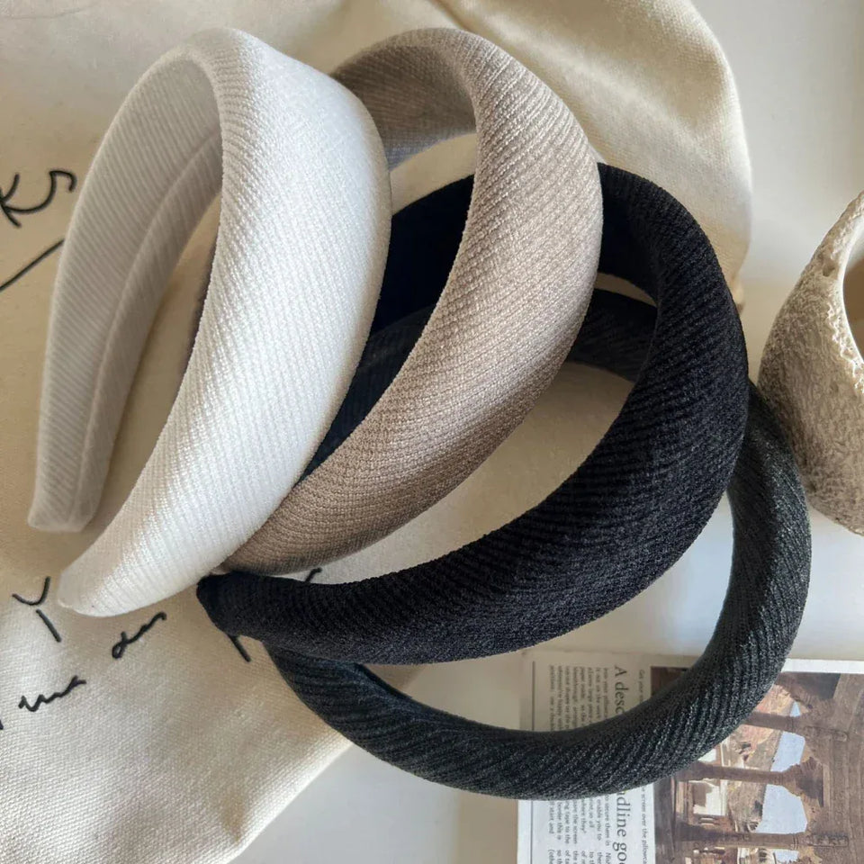 A collection of four padded headbands in various neutral colors (white, beige, black, dark gray), arranged on a neutral fabric surface with some decorative items. Secondary image