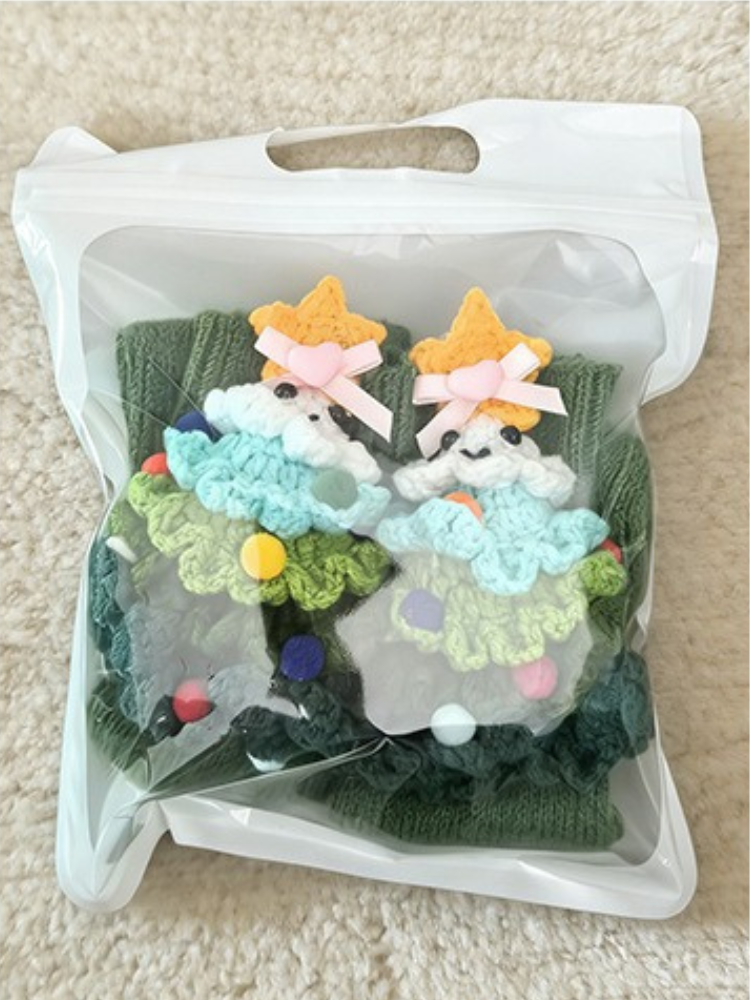 A pair of green ribbed knit crew socks with crocheted Christmas tree embellishments, packaged in a clear plastic bag.