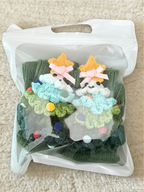 A pair of green ribbed knit crew socks with crocheted Christmas tree embellishments, packaged in a clear plastic bag.