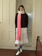 Full body shot of a woman wearing a brown sweatshirt, white ruffled skirt, and a long gradient scarf from cream to pink with fringe detail, standing in front of a bookshelf.