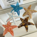 Five starfish hair clips (blue, yellow, white, orange, brown) arranged on a glass surface with a framed quote in the background