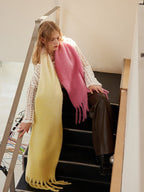 Woman wearing a two-tone yellow and pink fringed scarf draped over a white crochet top and brown wide-leg pants, sitting on stairs.