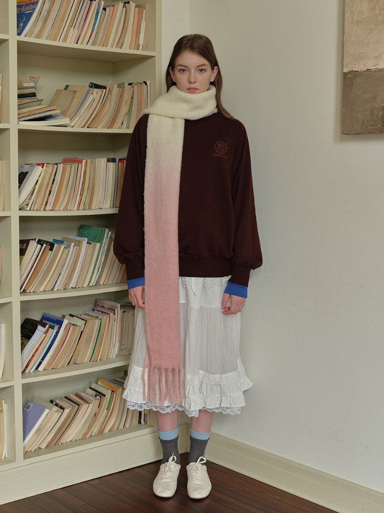 Woman wearing a brown sweatshirt, white ruffled skirt, and a long gradient scarf from cream to pink with fringe detail, standing in front of a bookshelf.