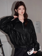 Woman adjusting the collar of her black faux leather jacket, revealing a grey crop top underneath.