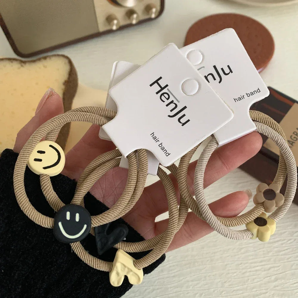 A hand holding three pairs of beige hair ties with various charms including smiley faces, hearts, and flowers.