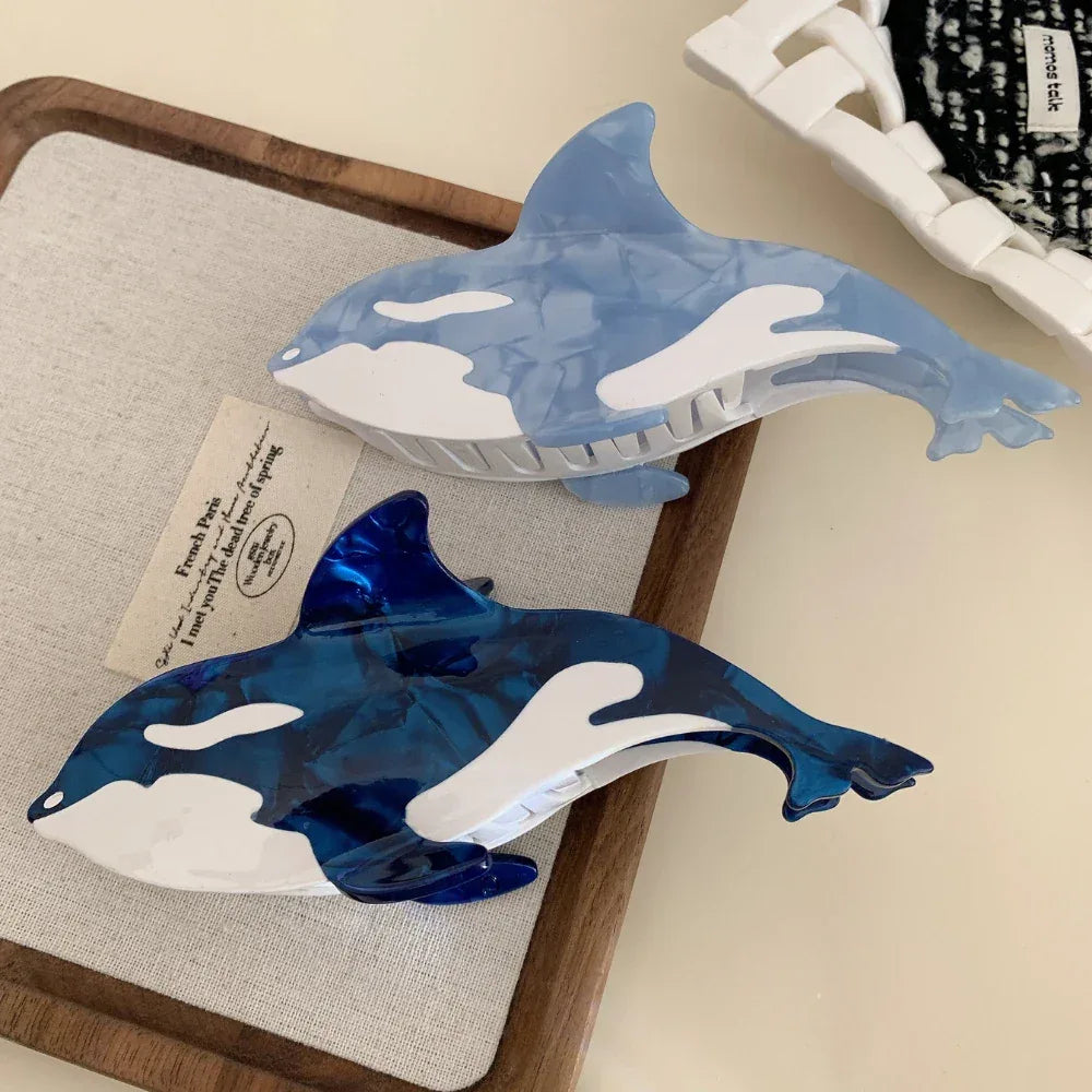 Two orca whale hair claw clips, one in a lighter blue and one in a darker blue, resting on a textured surface.