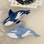 A close-up view of two orca whale hair claw clips, one dark blue and one light blue, on a neutral fabric background.