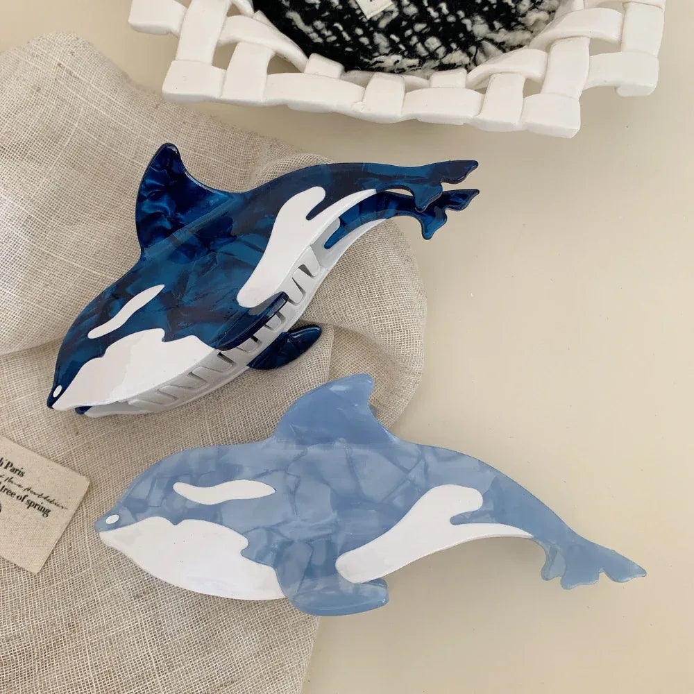 A close-up view of two orca whale hair claw clips, one dark blue and one light blue, on a neutral fabric background.