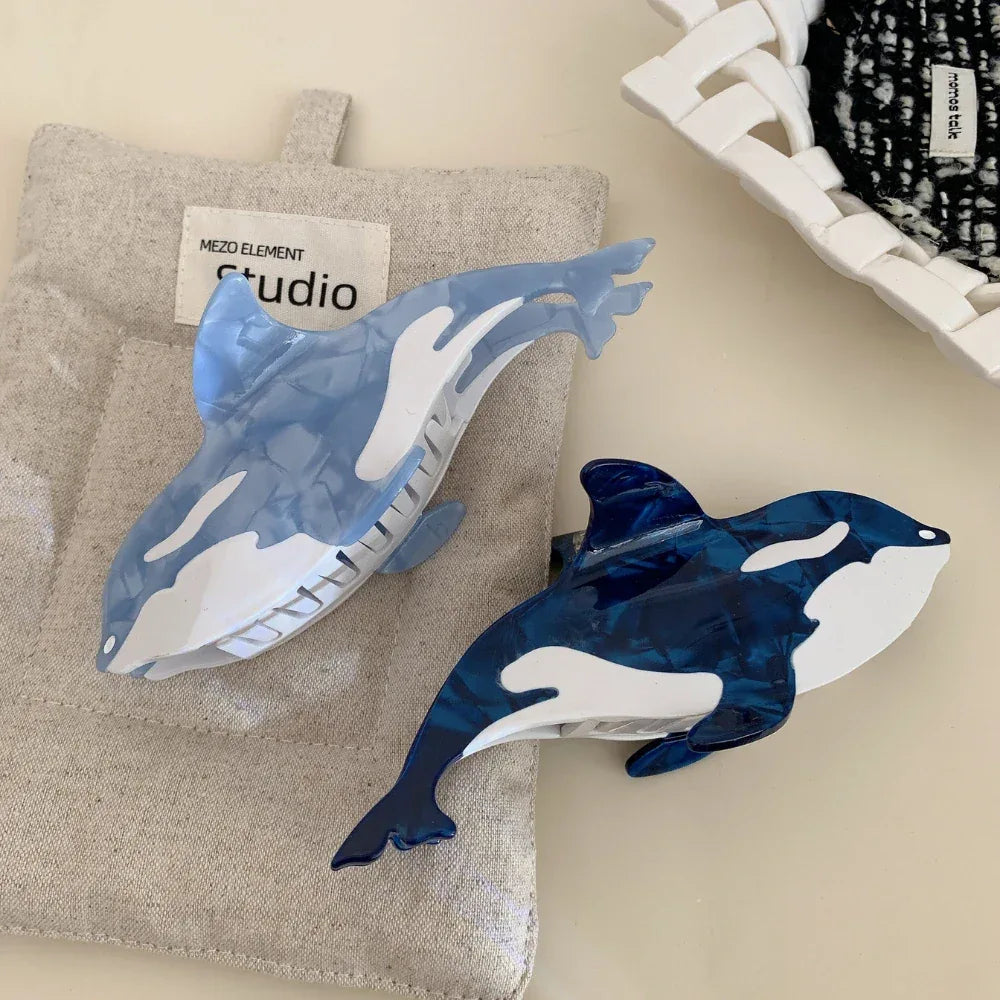 Two orca whale hair claw clips, one dark blue and one light blue, positioned on a light-colored linen-like fabric.