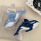 Two orca whale hair claw clips, one dark blue and one light blue, positioned on a light-colored linen-like fabric.