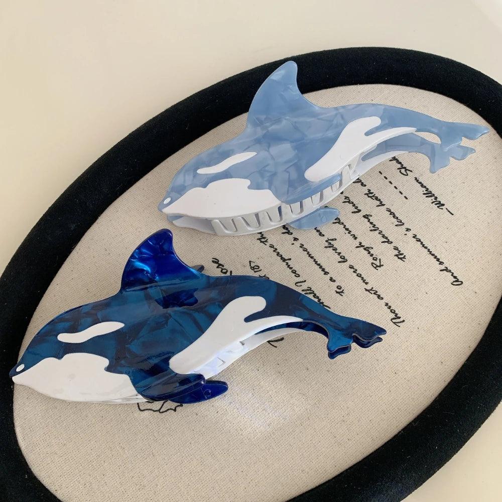 Two orca whale hair claw clips, one dark blue and one light blue, displayed on a decorative oval surface with text.