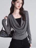Model holding a black handbag while wearing a grey long-sleeved top with a draped neckline.