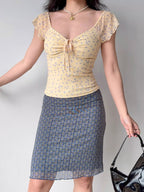 Full body shot of a woman wearing a yellow floral print mesh top and a patterned skirt.