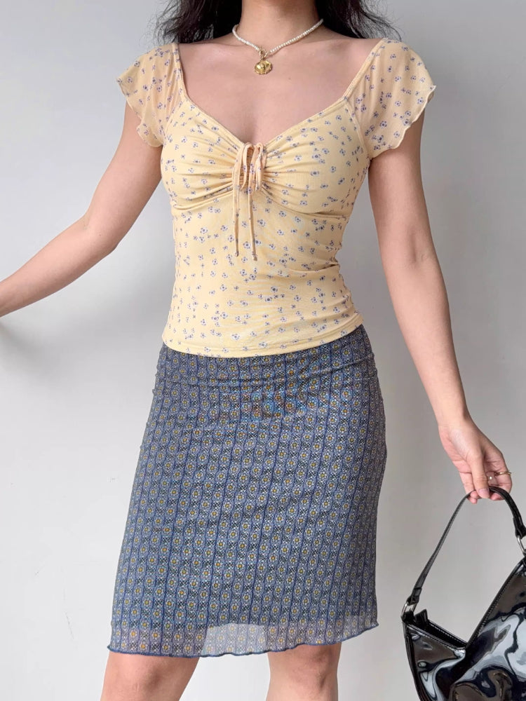 Full body shot of a woman wearing a yellow floral print mesh top and a patterned skirt.