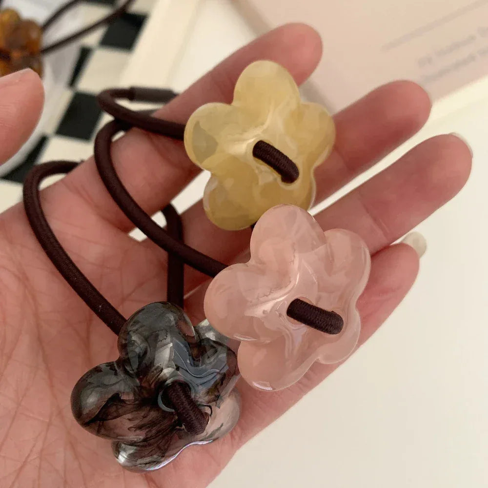 A hand holding three flower-shaped hair ties in yellow, pink, and grey marble patterns.