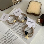 Three pairs of beige hair ties with charms are displayed on a newspaper with a radio and sponge in the background.