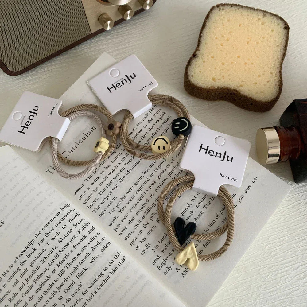 Three pairs of beige hair ties with charms are displayed on a newspaper with a radio and sponge in the background.