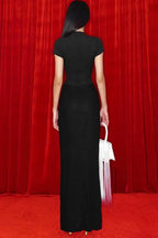 Back view of a model wearing a black long dress with a deep V-neckline and ruching on the skirt, holding a white bag with fringe details, standing in front of a red curtain.