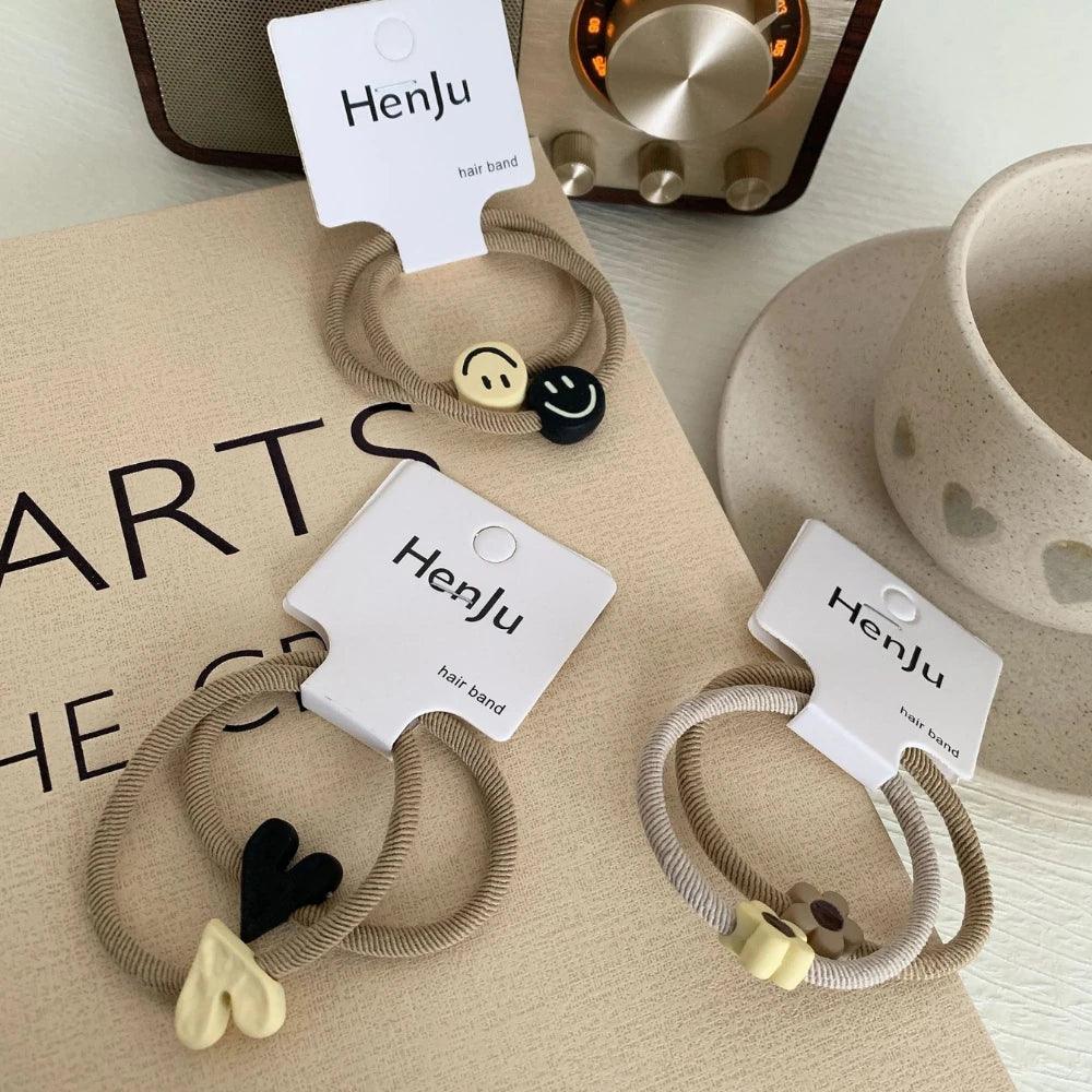 Four sets of beige hair ties with charms (smiley faces, hearts, flowers) are arranged on a table with a radio and cup.