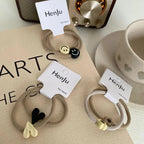 Four sets of beige hair ties with charms (smiley faces, hearts, flowers) are arranged on a table with a radio and cup.