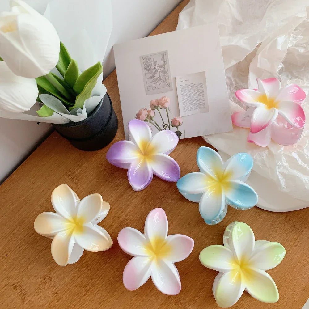 A collection of plumeria flower hair clips in various pastel colors, including pink, purple, blue, yellow, and green, arranged on a wooden surface with decorative elements.