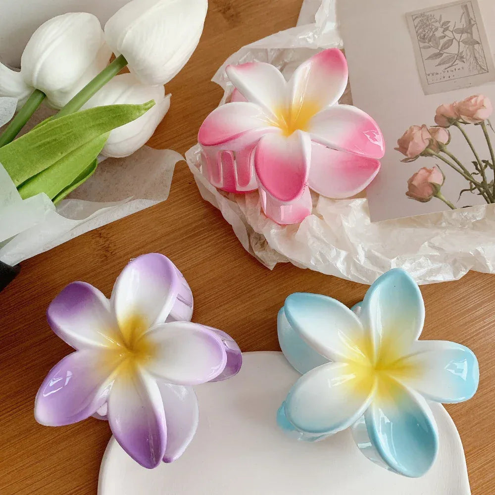Close-up of purple, blue, and pink plumeria flower hair clips on a white dish, with decorative paper and flowers in the background.