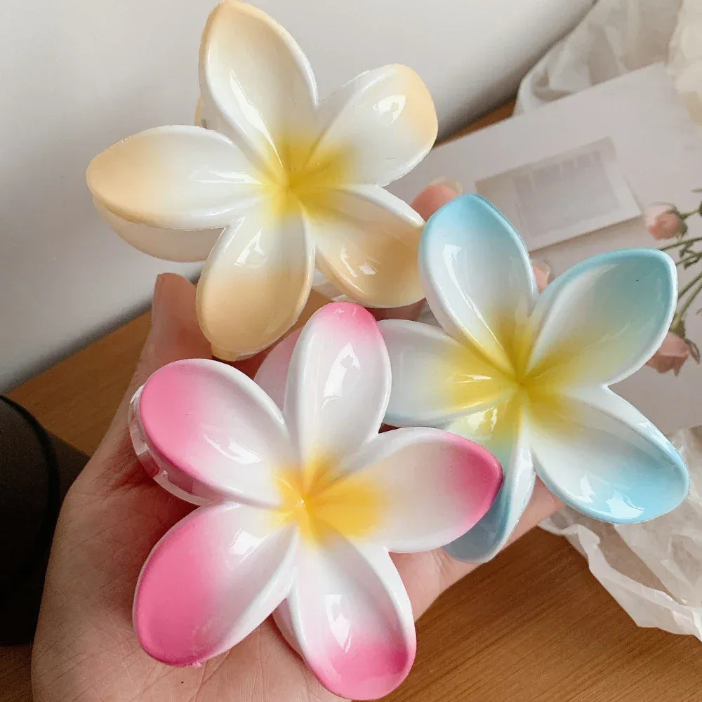 A hand holding three plumeria flower hair clips in pink, blue, and yellow, showcasing their vibrant colors and glossy texture.