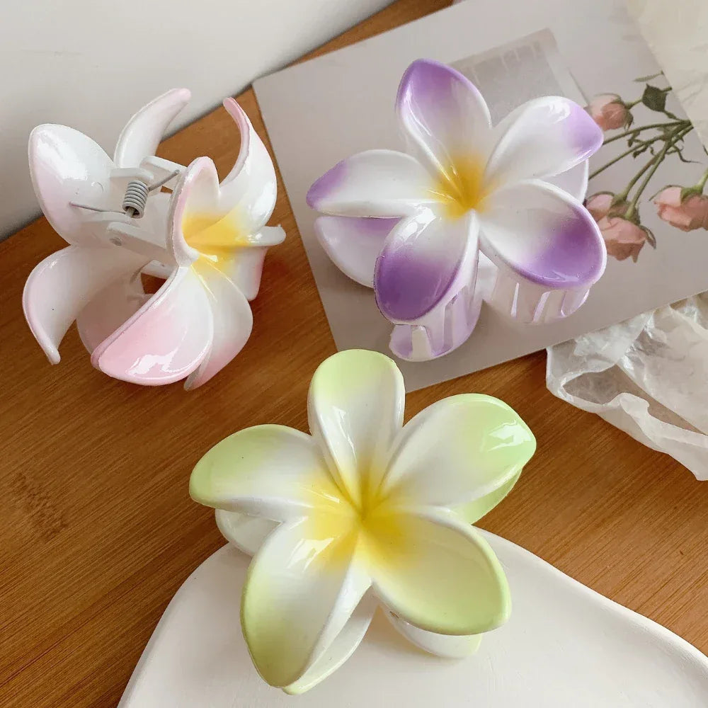 A selection of plumeria flower hair clips in pink, purple, and green, displayed on a white dish and a wooden surface.