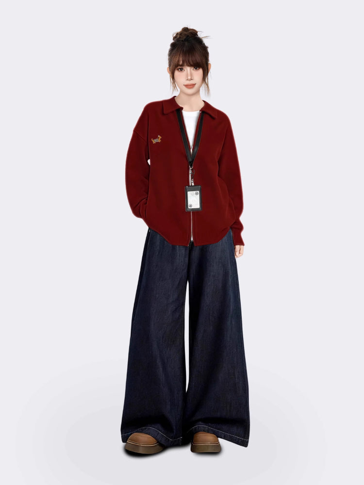 Full-body shot of a woman wearing a maroon zip-up knit cardigan, white t-shirt, and dark wash wide-leg jeans, standing against a white background.