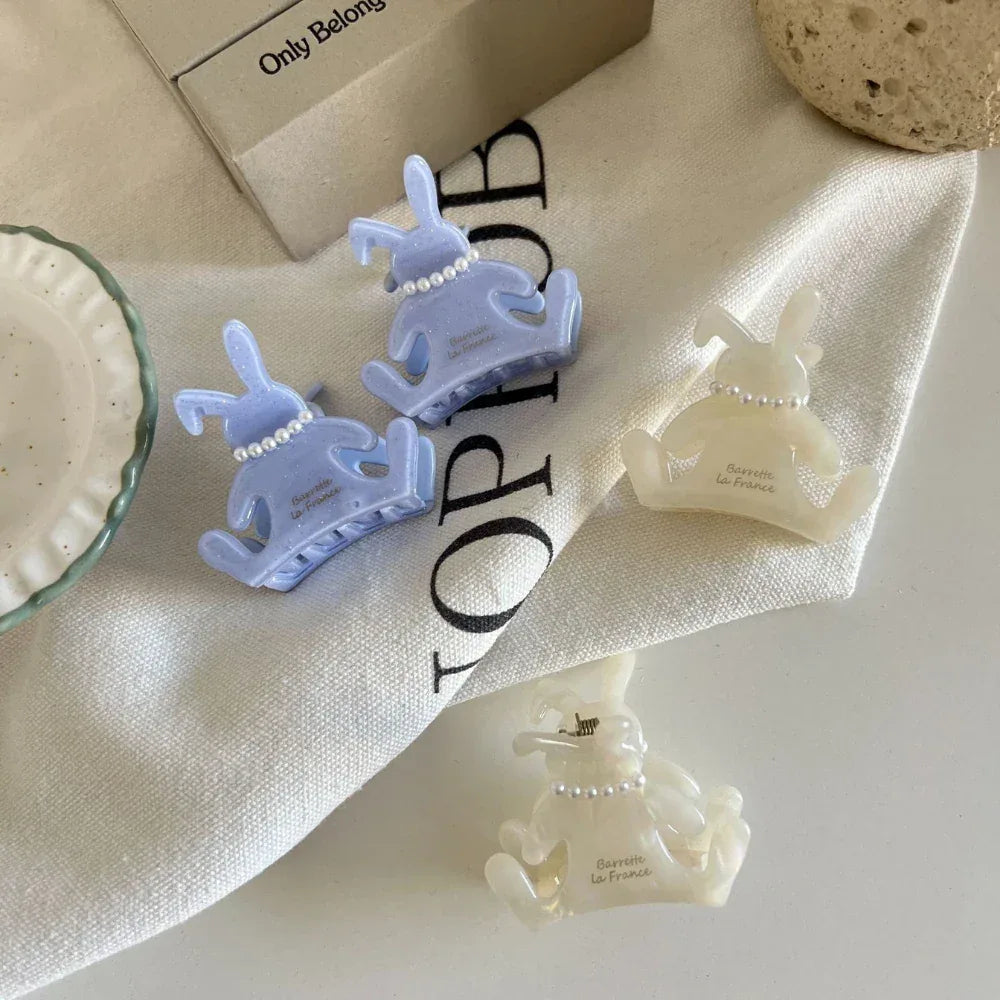 Four bunny-shaped hair claw clips in white and light purple, arranged on a white cloth with text.