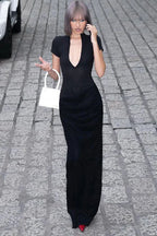 Model wearing a black long dress with a deep V-neckline and ruching on the skirt, holding a white bag with fringe details, walking on a cobblestone street.