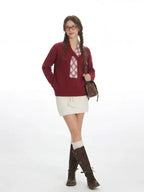 Woman stands with hands in pockets, wearing a burgundy v-neck sweater with a white plaid detail, a white textured mini skirt, white knee-high socks, and brown buckled boots, carrying a brown crossbody bag.
