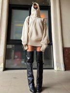 Woman in a beige oversized knit hooded sweater dress and black thigh-high boots, standing straight.
