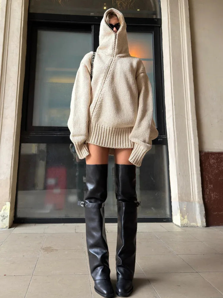 Woman in a beige oversized knit hooded sweater dress and black thigh-high boots, standing straight.