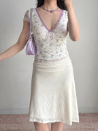 Woman in a white floral print top and white textured skirt, holding a purple handbag and looking to the side.