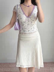 Woman in a white floral print top and white textured skirt, holding a purple handbag and looking to the side.