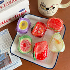 A collection of fruit-shaped hair clips arranged on a plate, including a strawberry, watermelon, grapefruit, tomato, fig, and pear.
