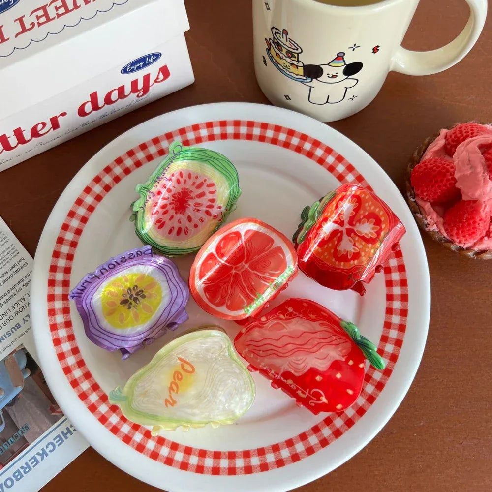 A platter of assorted fruit-shaped hair clips, including a strawberry, watermelon, grapefruit, tomato, fig, and pear, arranged with a checkered napkin and a mug.