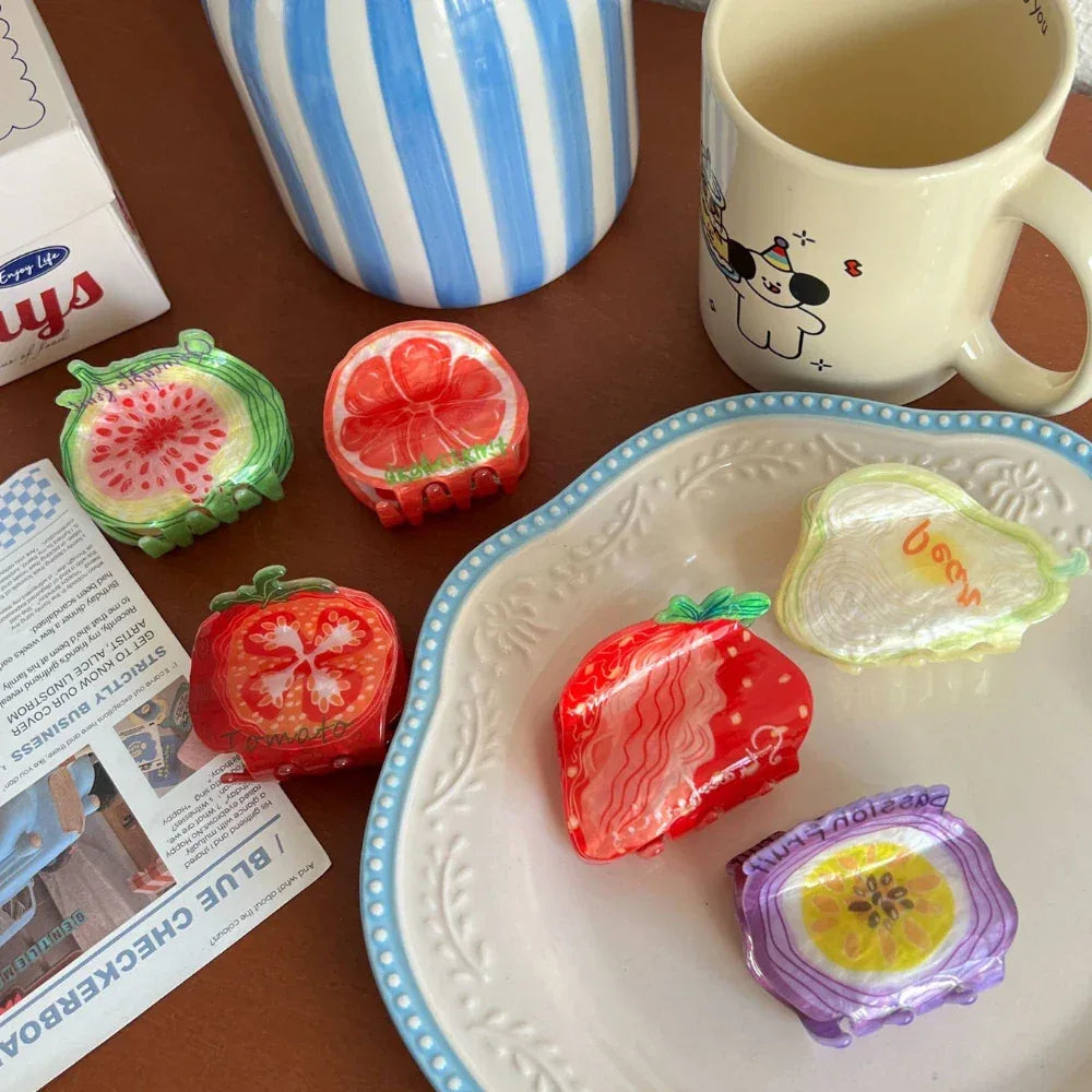 A top-down view of various fruit-shaped hair clips displayed on a table next to a mug and some packaging.
