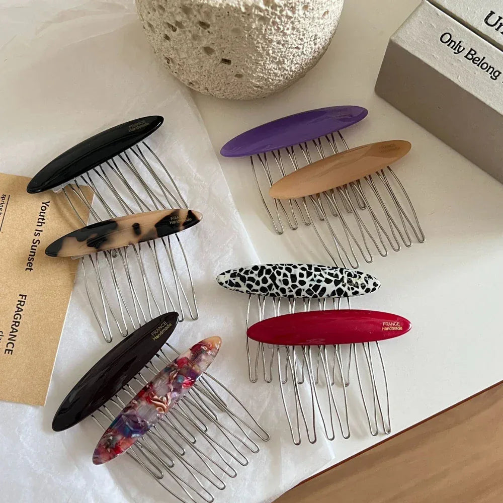 A variety of hair combs with oval decorative tops in different colors and patterns are laid out on a white surface, with some paper tags and a textured pot in the background.