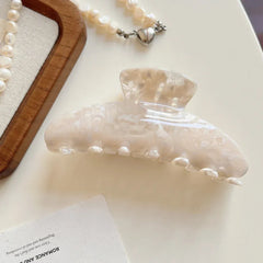 A single cream-colored marbled acetate claw clip is shown on a white surface next to a card and a pearl bracelet.