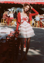 Woman in a white dress with red trim and bows, walking outdoors near a restaurant.