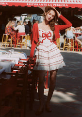 Woman in a white dress with red trim and bows, walking outdoors near a restaurant.