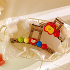 A transparent hair claw clip with a red apple charm and colorful beads, resting in a white basket lined with tissue paper.