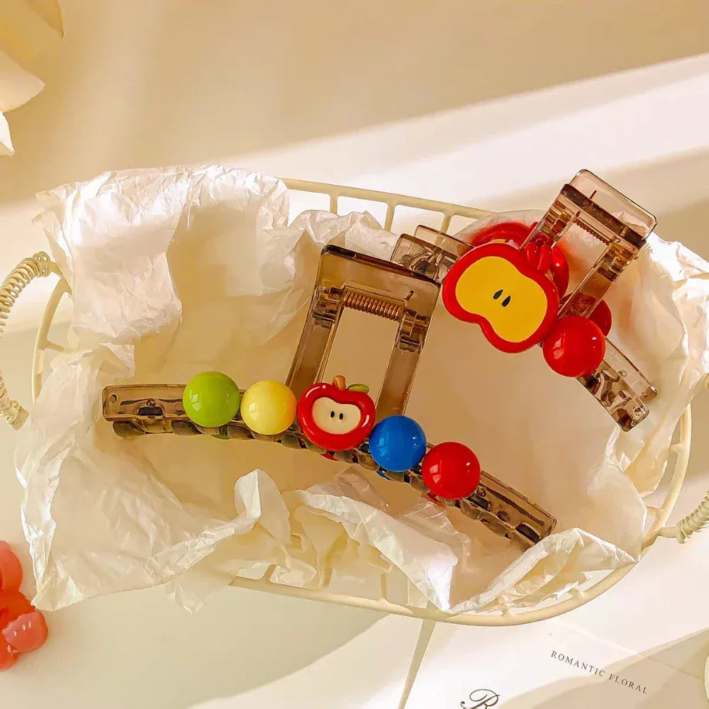 A transparent hair claw clip with a red apple charm and colorful beads, resting in a white basket lined with tissue paper.