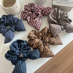 Collection of plaid scrunchies with bow details in various colors including navy, burgundy, brown, and tan.