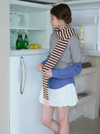 Model reaching into an open refrigerator, wearing a layered outfit with a grey cropped sweater, blue checkered shirt, white ruffled skirt, and striped scarf.