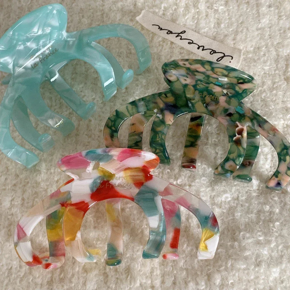 Three marbled acetate claw hair clips are displayed on a white textured surface: a multi-colored one, a light blue one, and a green one. A white ribbon with text is in the background.