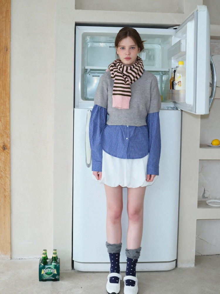 Full-body shot of a model in a layered outfit featuring a grey cropped sweater, blue checkered shirt, white ruffled skirt, striped scarf, and white Mary Jane shoes, standing in front of a refrigerator.